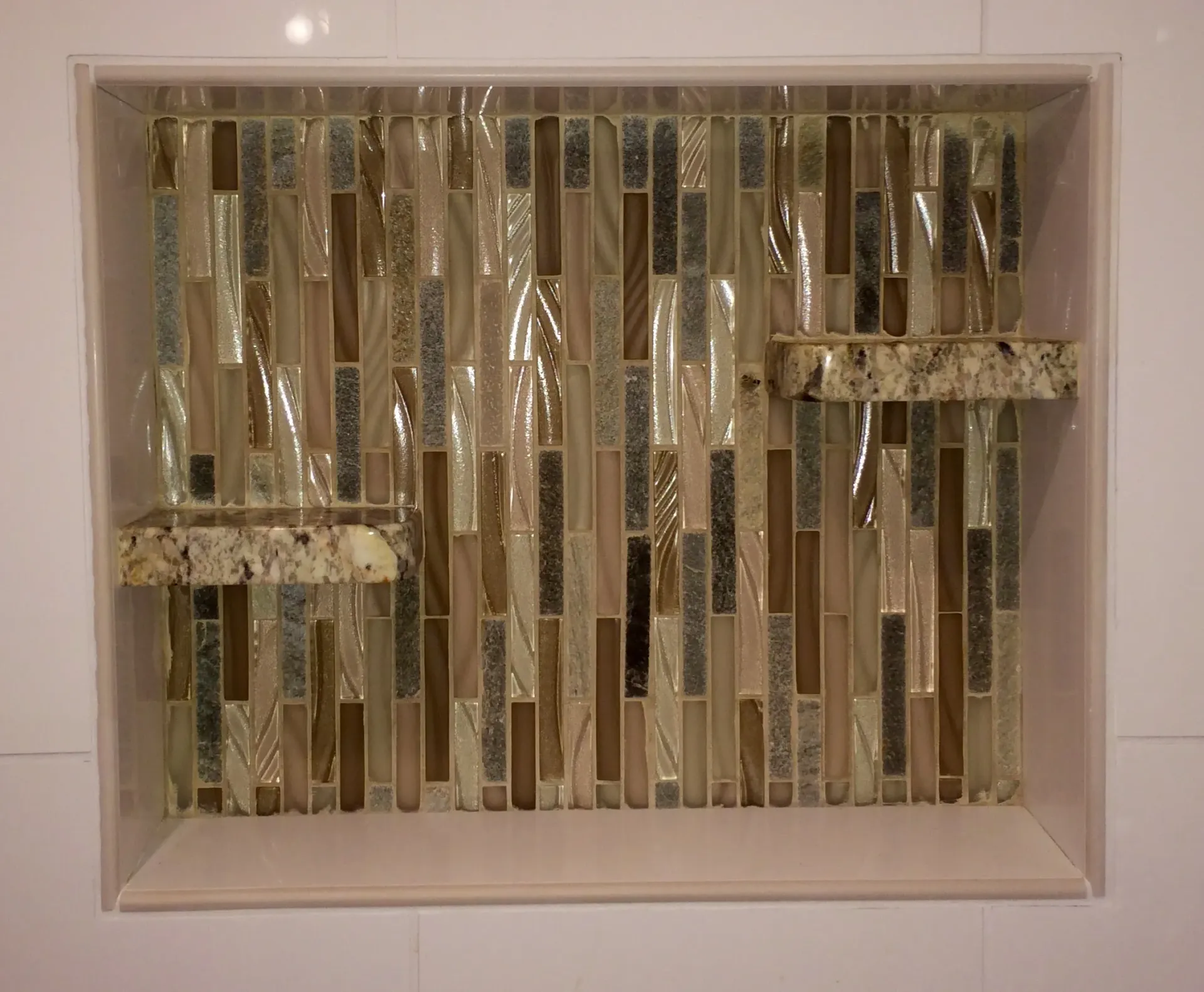 A recessed shower niche with a decorative vertical tile mosaic, two granite corner shelves, and a large bottom ledge.