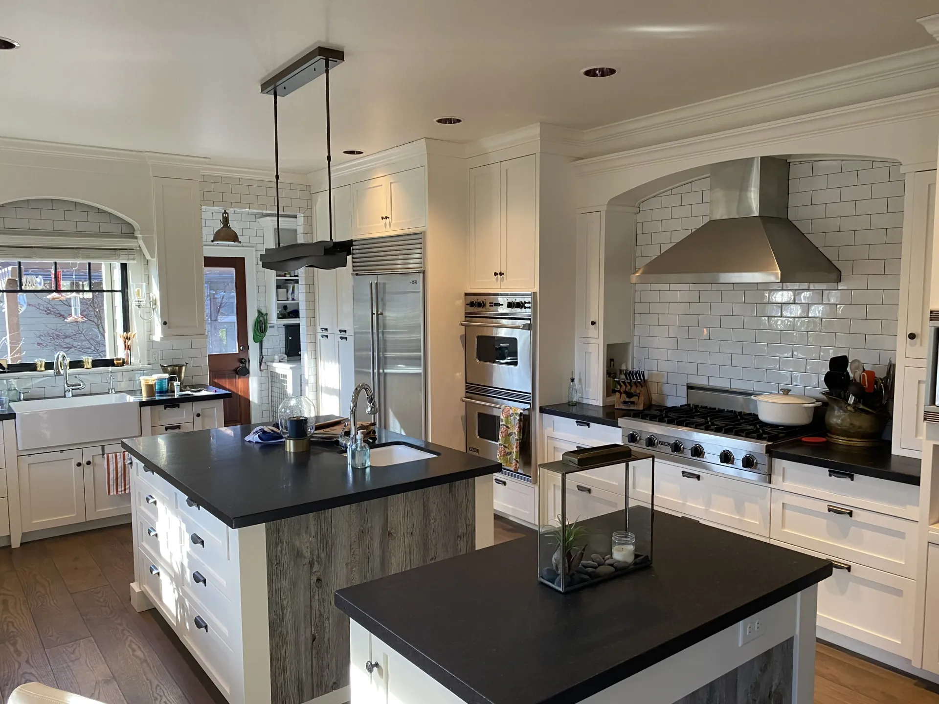 A bright kitchen featuring two dark-topped islands, white cabinets, stainless steel appliances, and a tiled backsplash.