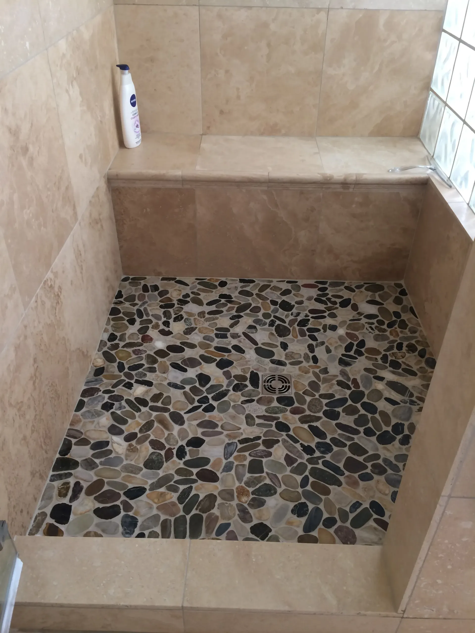 A shower stall featuring tan stone wall tiles, a stone bench with a bottle of lotion, and a dark pebble mosaic floor.