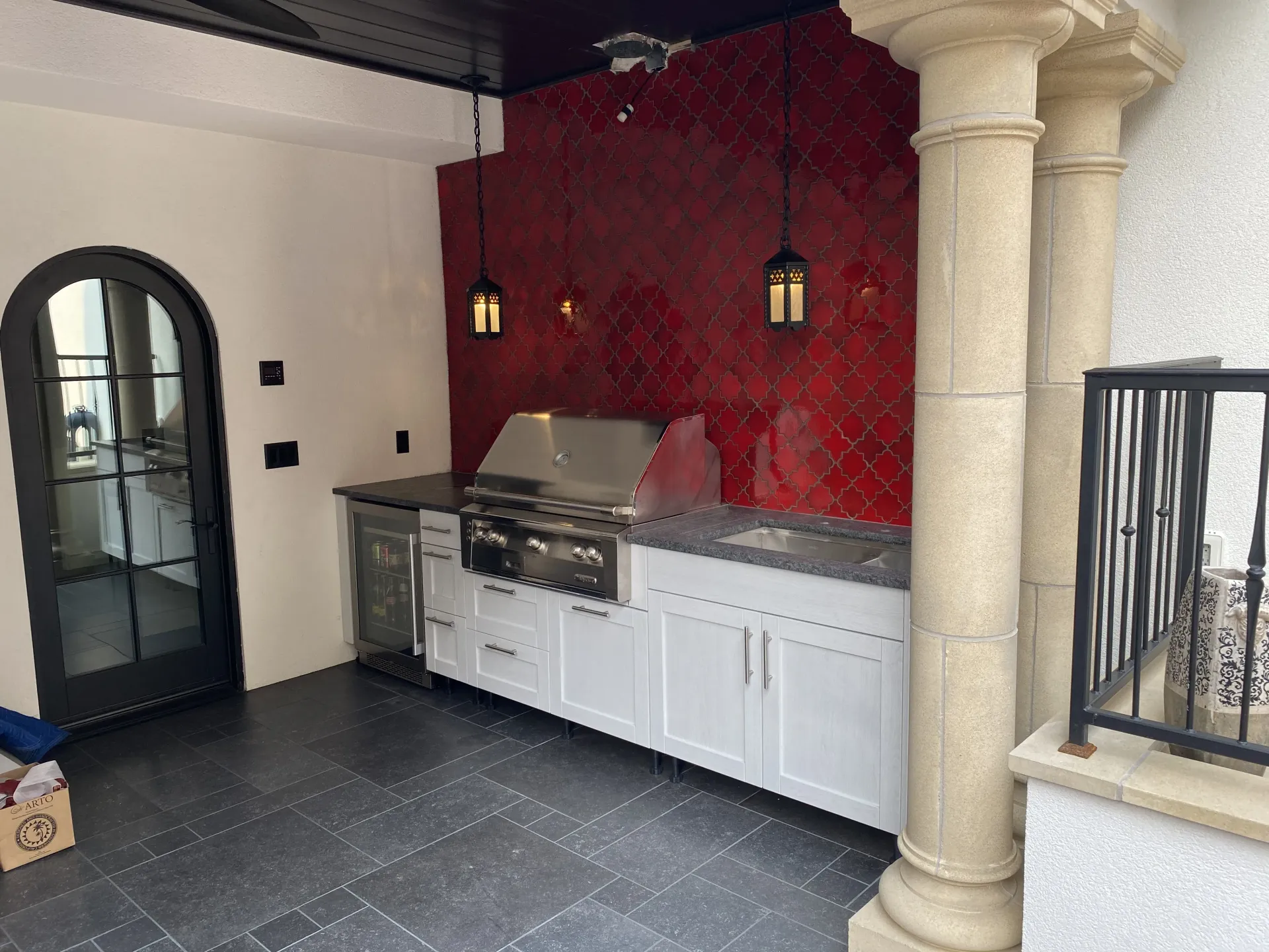An outdoor kitchen with white cabinets, a grill, and a vibrant red tiled backsplash, next to a stone pillar and doorway.