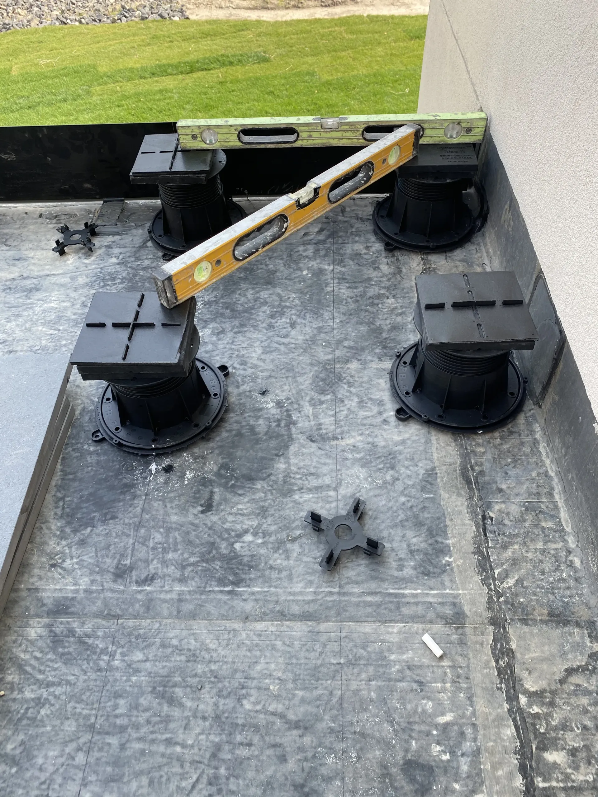Four adjustable black deck pedestals are positioned on a grey flat roof, with spirit levels resting across them.