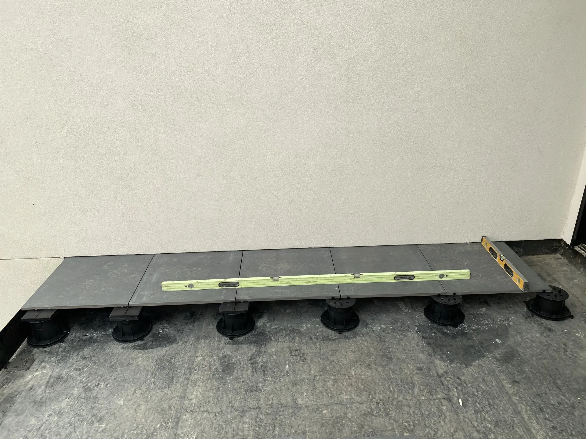 A row of grey floor tiles laid on adjustable plastic pedestals against an exterior wall, with a spirit level on top.