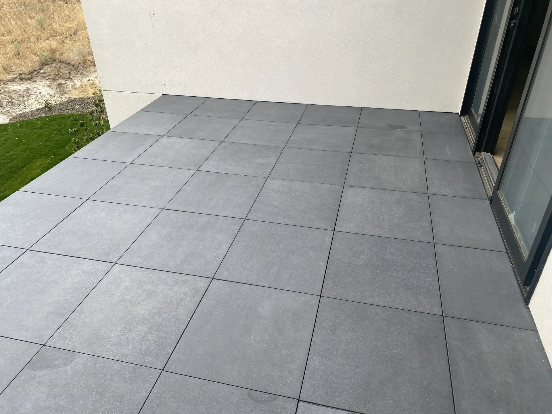 A grey, square-tiled patio floor adjacent to an exterior white wall and a glass doorway.