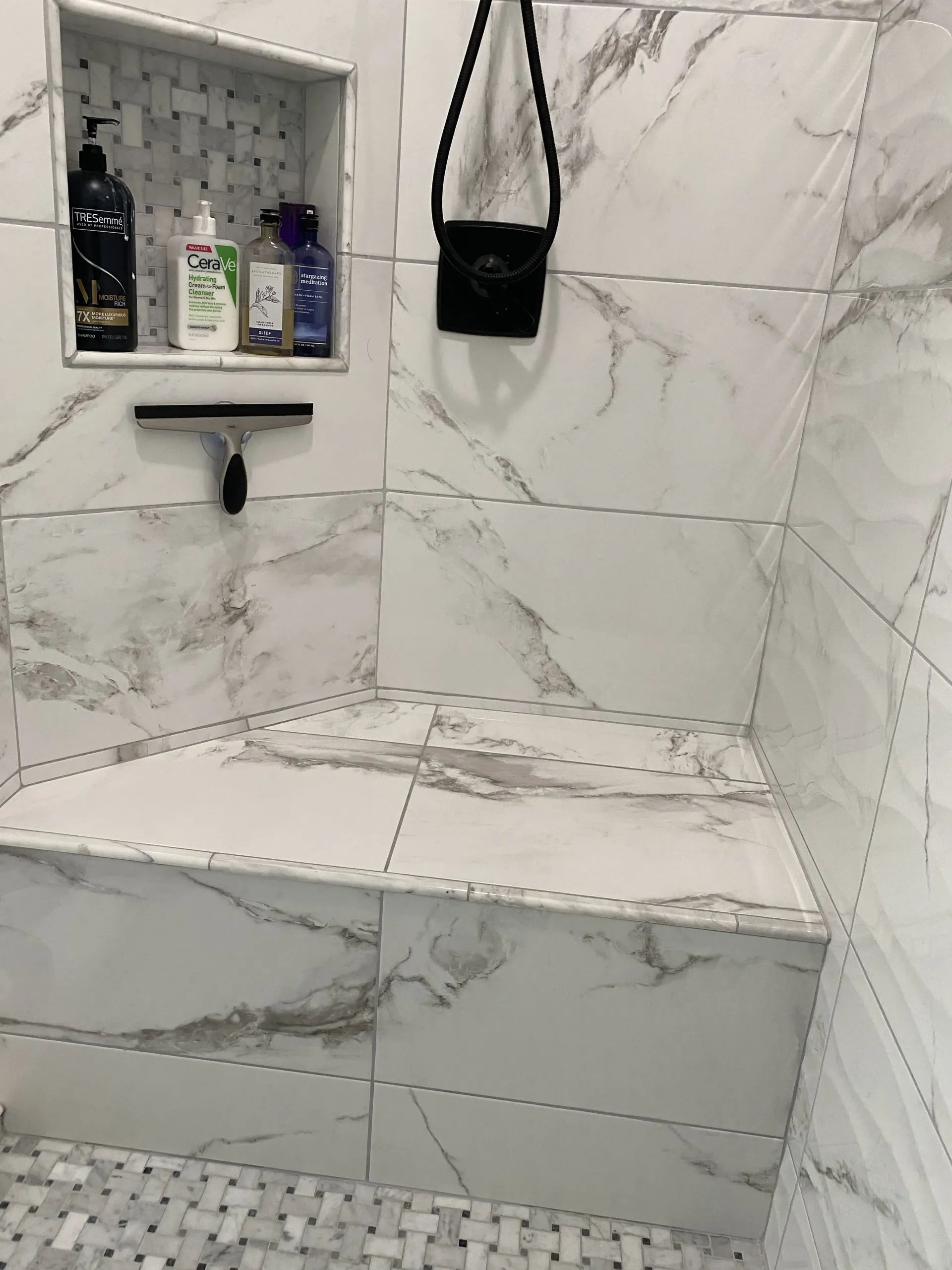 A shower with white marble-patterned tile, a built-in corner bench, a recessed shelf containing toiletries, and a squeegee.