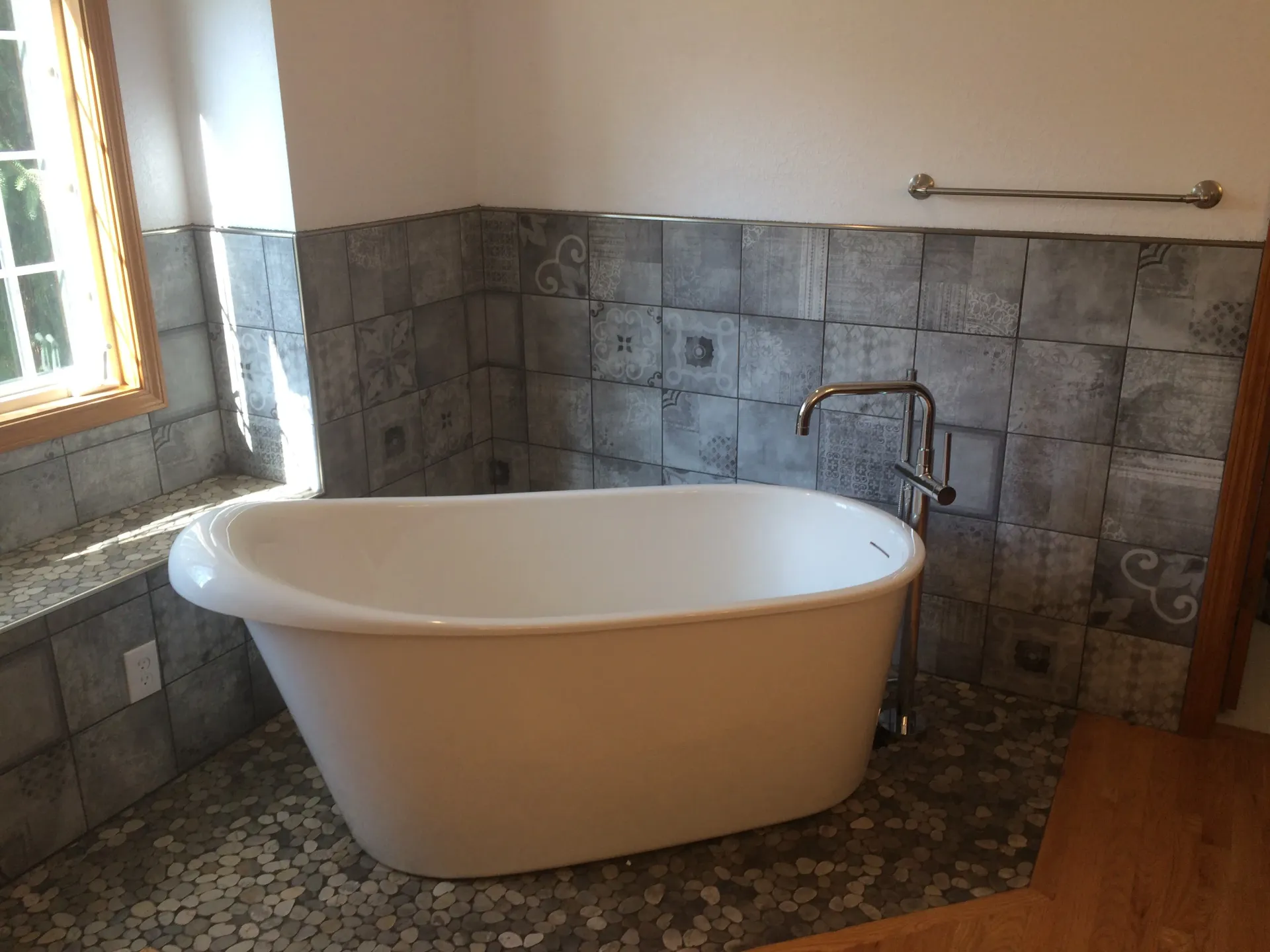 A white freestanding bathtub stands next to a window in a bathroom with gray tiled walls and a pebble floor.