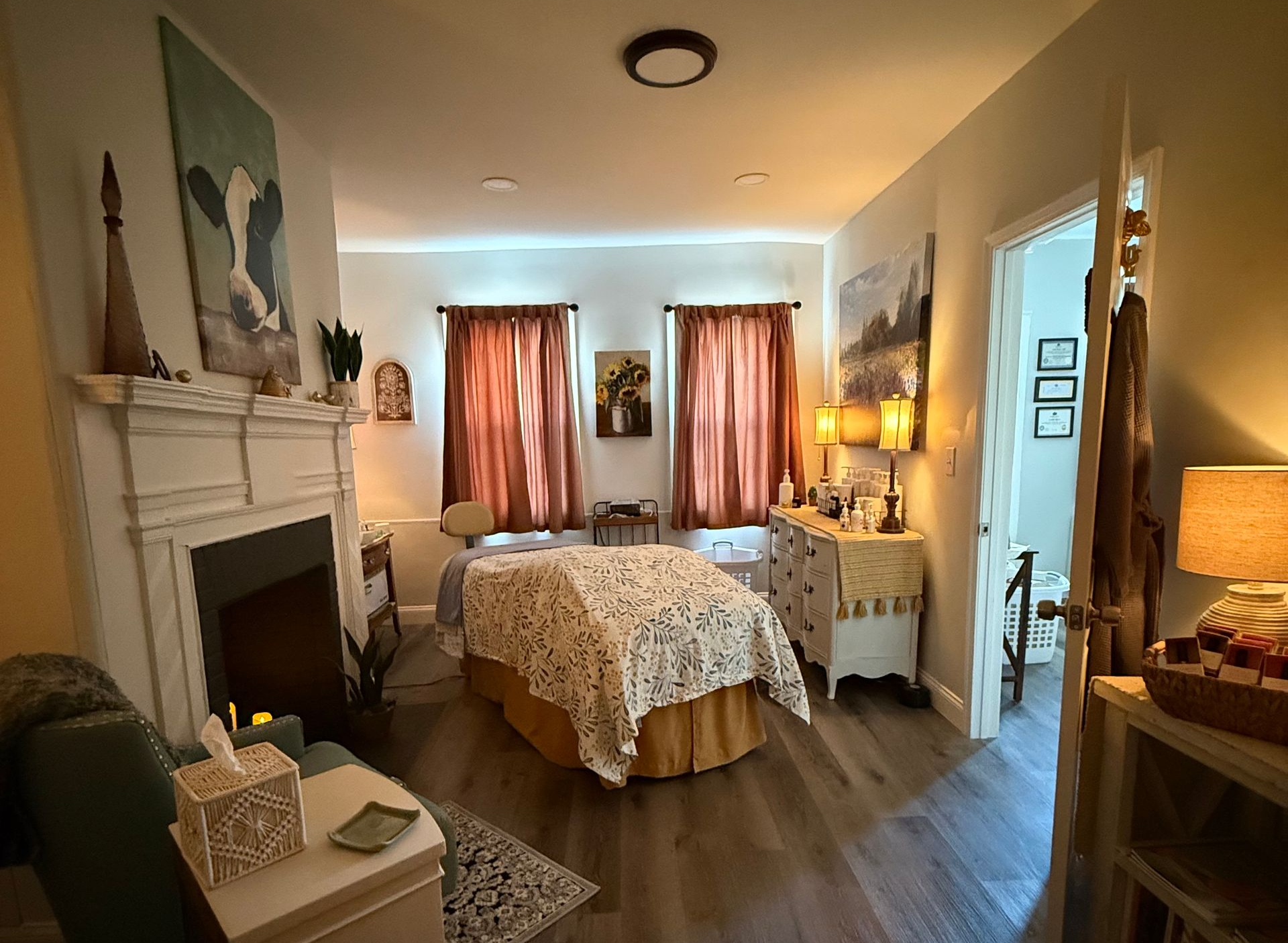 Cozy bedroom with two beds, red curtains, and artwork over a fireplace. A door opens to another room.