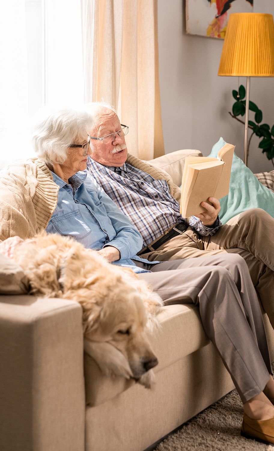 An elderly couple is sitting on a couch with a dog and reading a book.