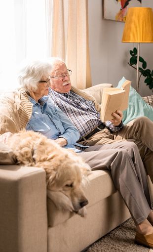 An elderly couple is sitting on a couch with a dog and reading a book.