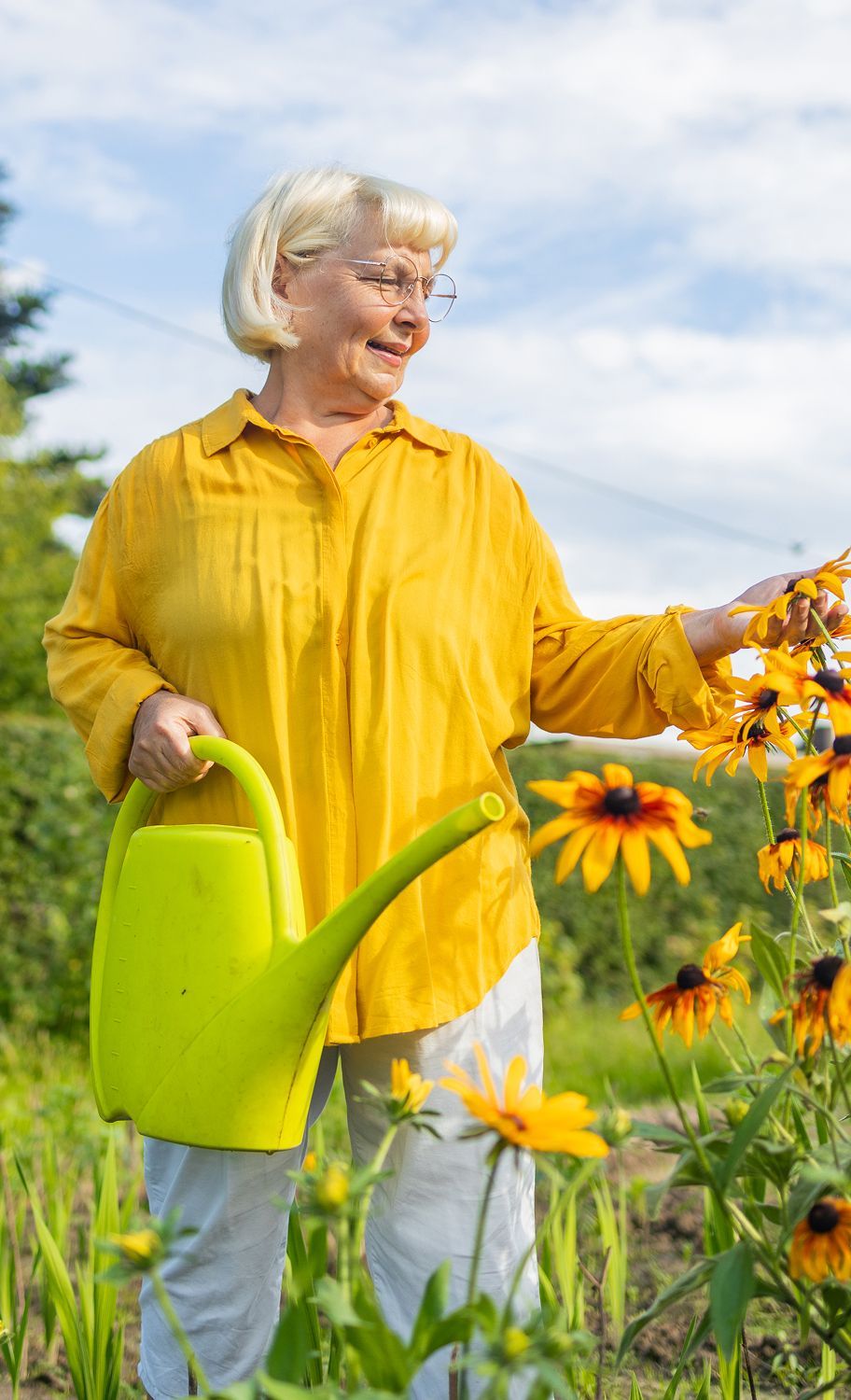An elderly woman is watering flowers in a garden with a watering can.