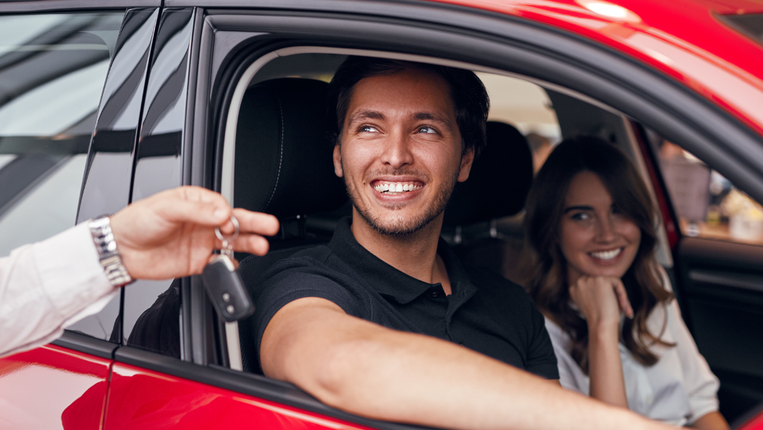 A man and woman are sitting in a red car while a man gives them the keys.