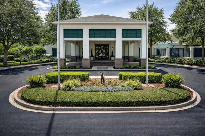 White building with columns, surrounded by greenery and circular driveway. Blue sky.