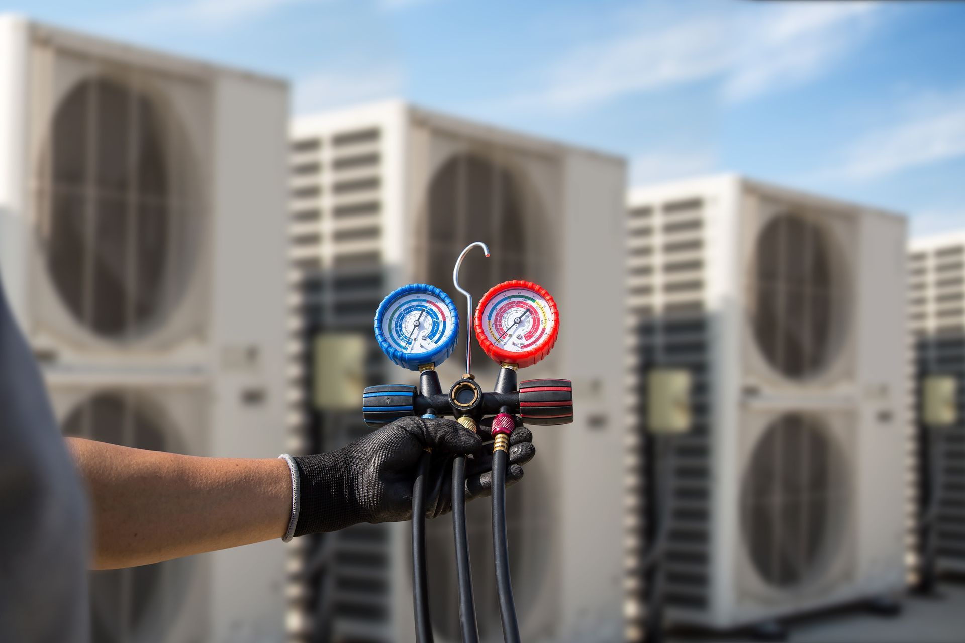 Gloved hand holding HVAC gauges in front of rooftop air conditioning units