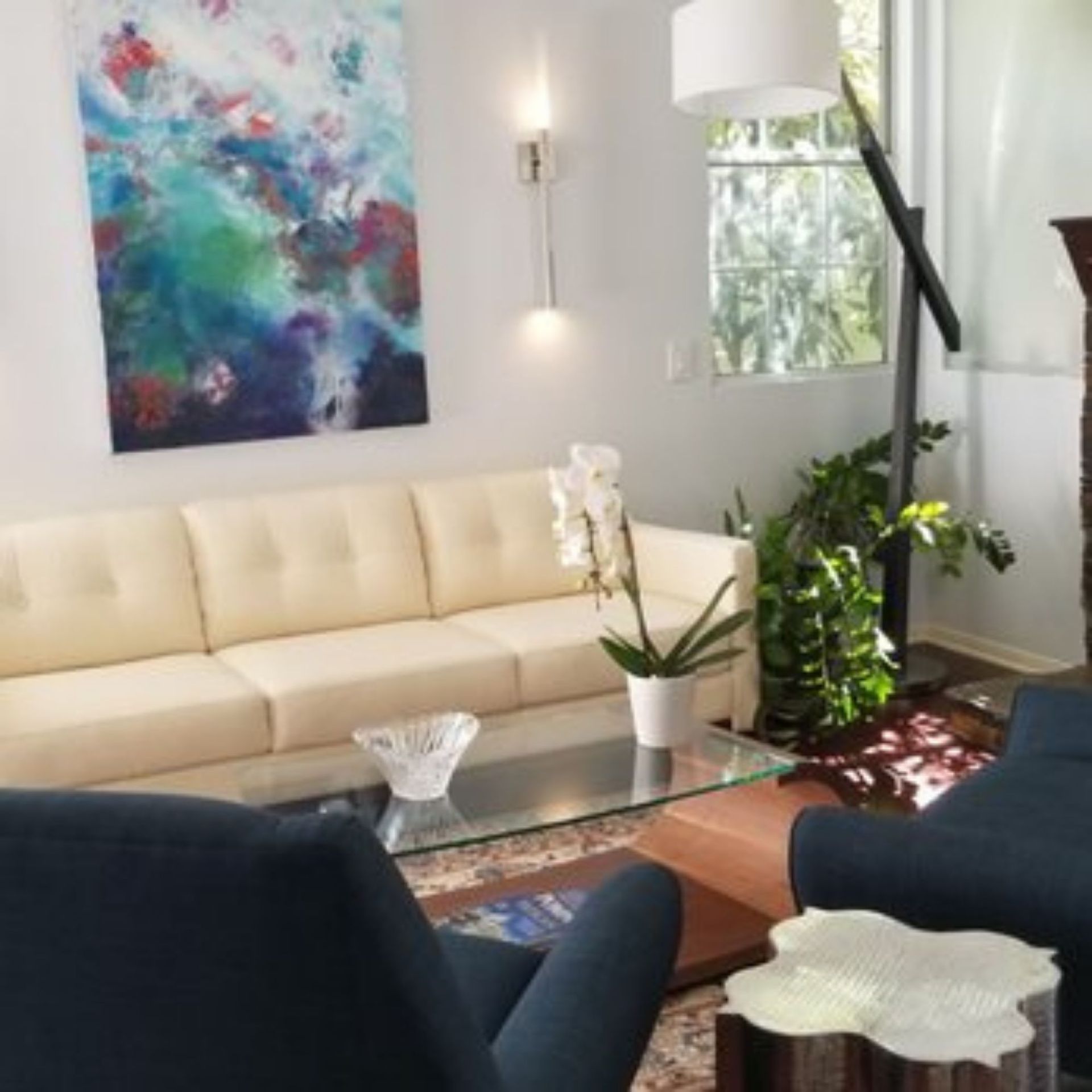 The right color - Living room with cream sofa, blue chairs, colorful artwork, glass coffee table, and plants.