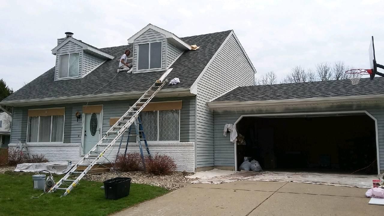 House being worked on; ladder on roof; two people; blue siding; garage open; concrete driveway.
