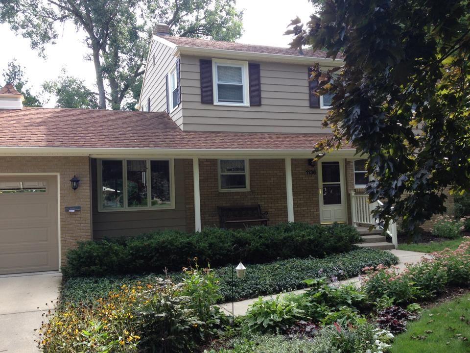 Two-story beige house with brown roof, surrounded by landscaping and trees.