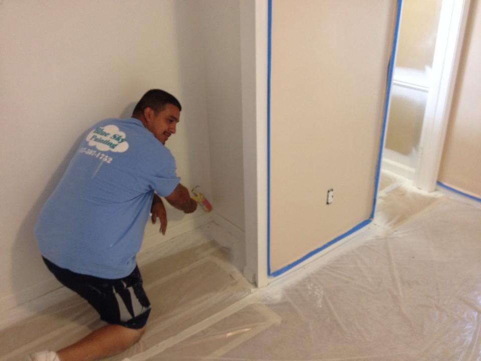 Painter using a brush to paint baseboard in room, masking tape on walls, plastic on the floor.