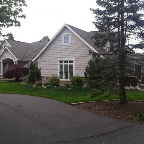 House with gray siding, dark roof, arched entryway, and lush landscaping on a curved driveway.