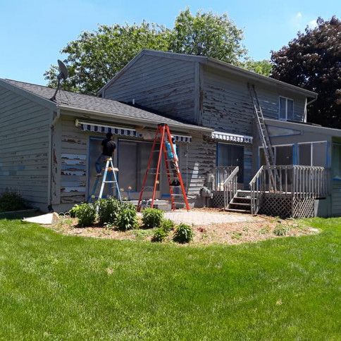 Two people on ladders installing an awning on a house's exterior with peeling paint.