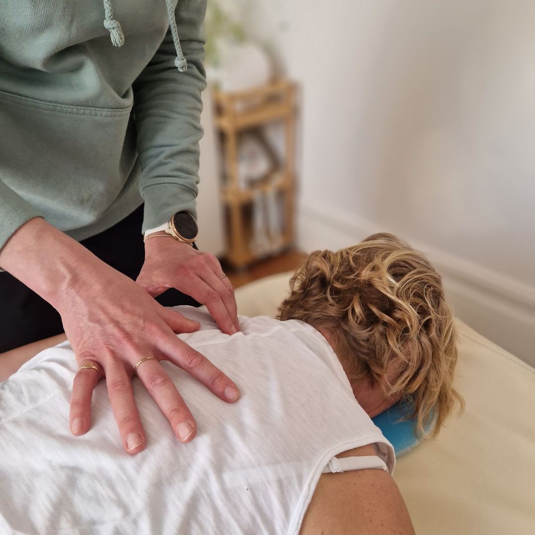 A patient sits on a treatment table with a female osteopath standing behind, with a hand on his head and shoulder