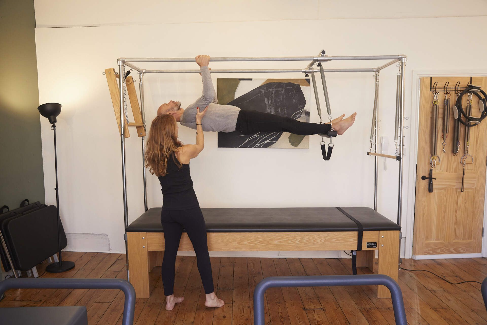 A Pilates teacher stands next to a Tower machine, assisting a student who is holding on the to the Tower in a horizontal pose