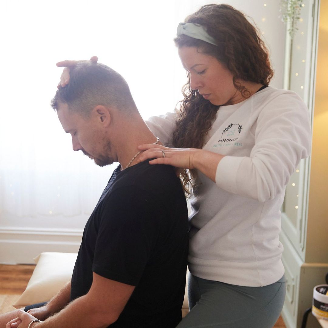 A patient sits on a treatment table with a female osteopath standing behind, with a hand on his head and shoulder