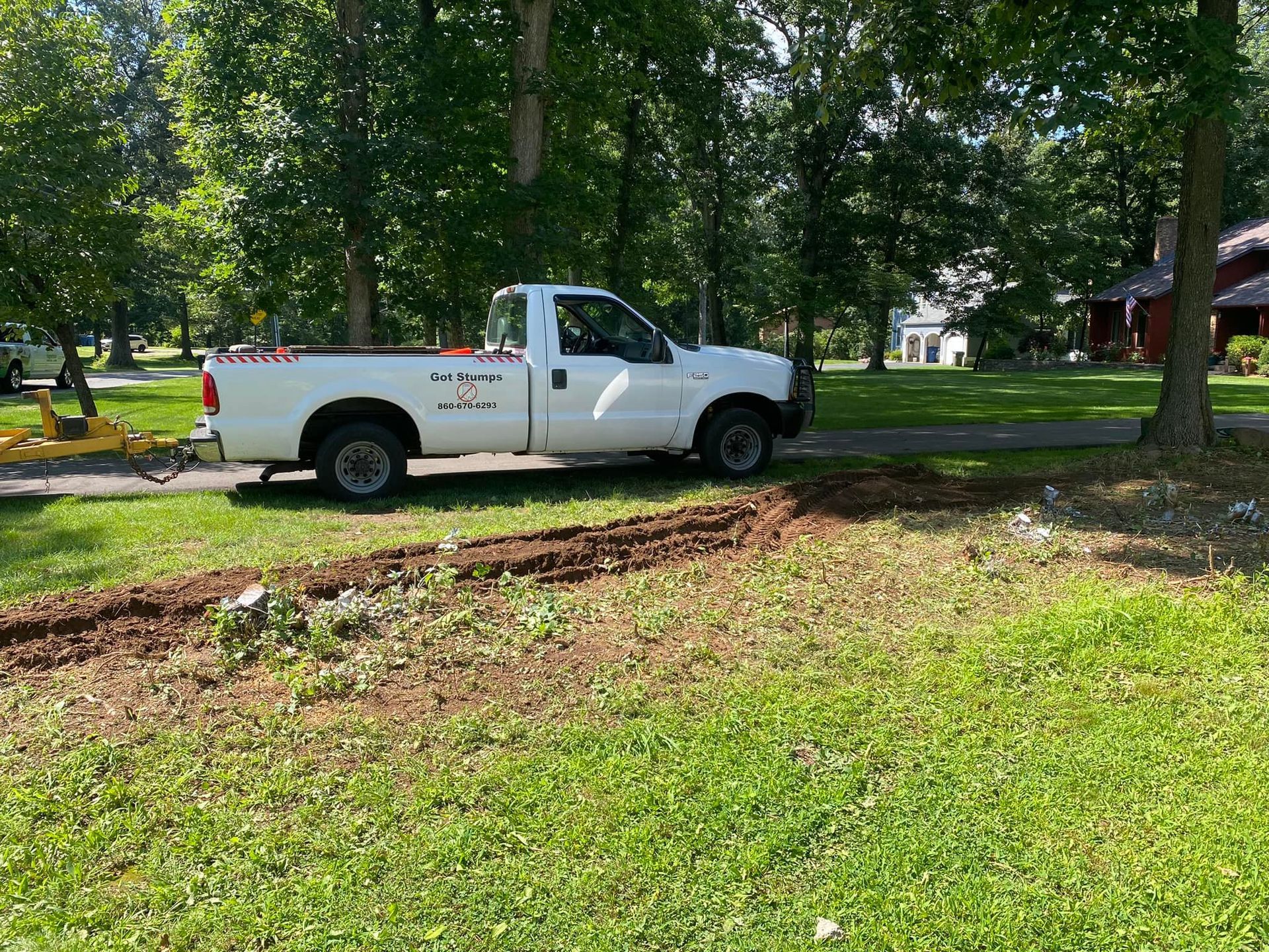 A white pickup truck parked on a grassy lawn next to a fresh patch of turned earth in a wooded, residential setting.