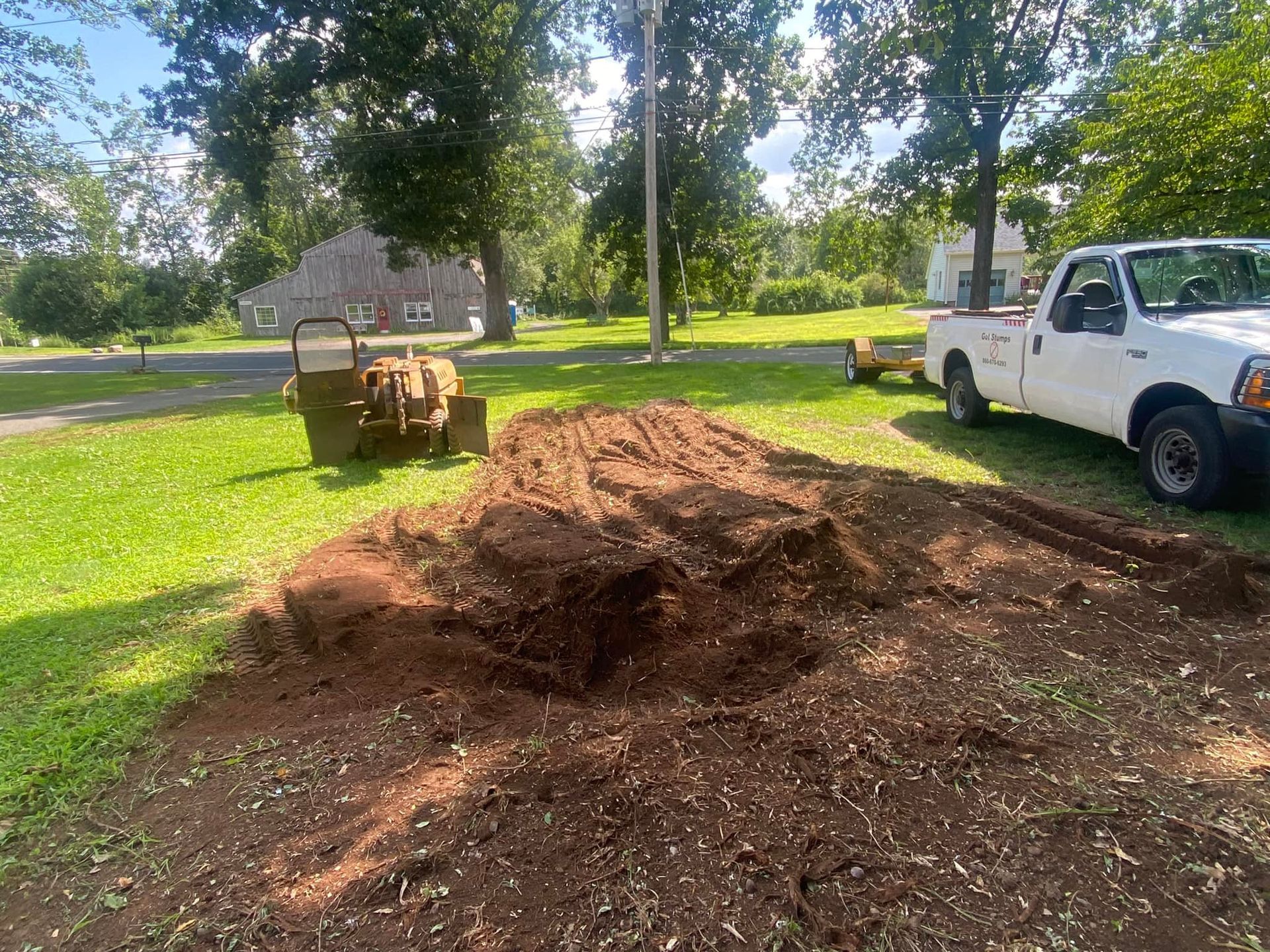 A white pickup truck parked next to a patch of freshly cleared, dark brown soil in a grassy, tree-lined yard.