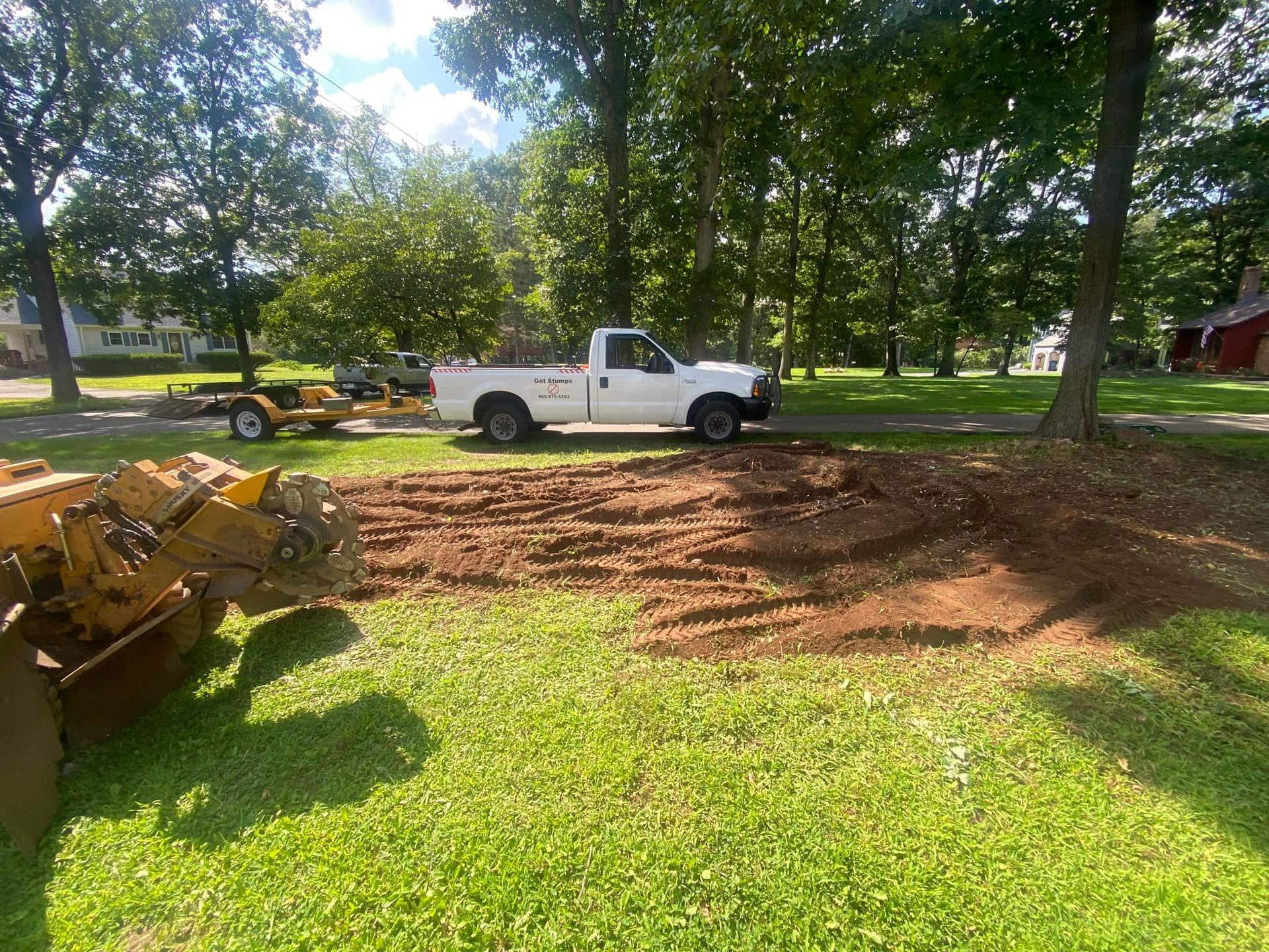 A yellow stump grinder sits on a grassy yard next to a patch of tilled soil, with a white pickup truck parked nearby.