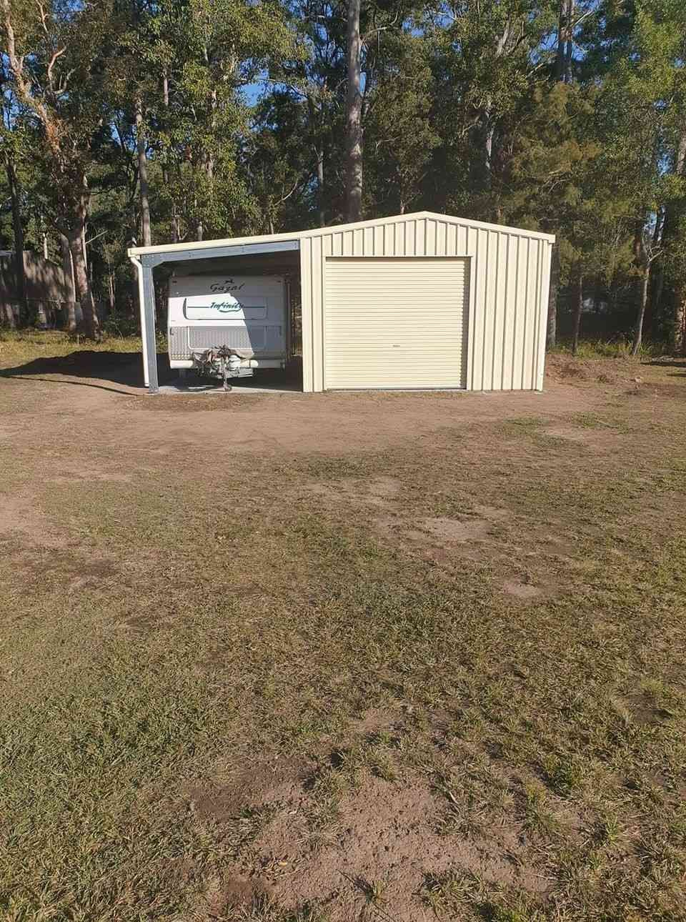 A Light Beige Metal Shed With a Recreational Vehicle Inside, Set in a Grassy Field With Trees in the Background — North Coast Sheds & Garages in Maclean, NSW
