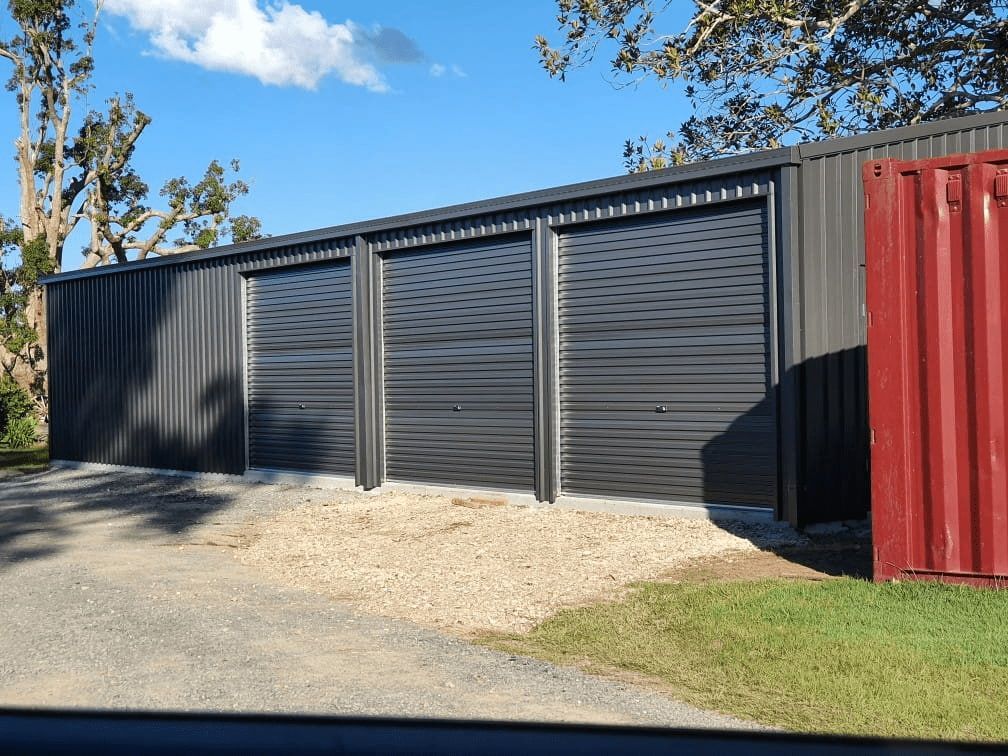 A Dark Gray Metal Shed With Three Garage Doors Sits on a Gravel Pad, Next to a Red Shipping Container, With a Blue Sky Overhead — North Coast Sheds & Garages in Coraki, NSW