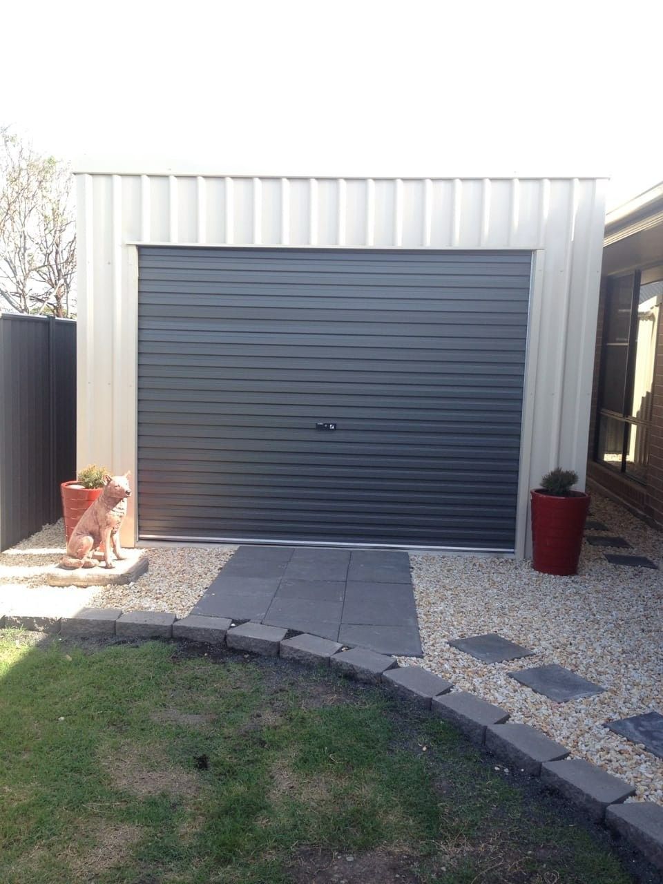 A Gray Garage With a Closed Roller Door, Flanked by Planters and a Stone Pathway in a Backyard Setting — North Coast Sheds & Garages in Brunswick Heads, NSW