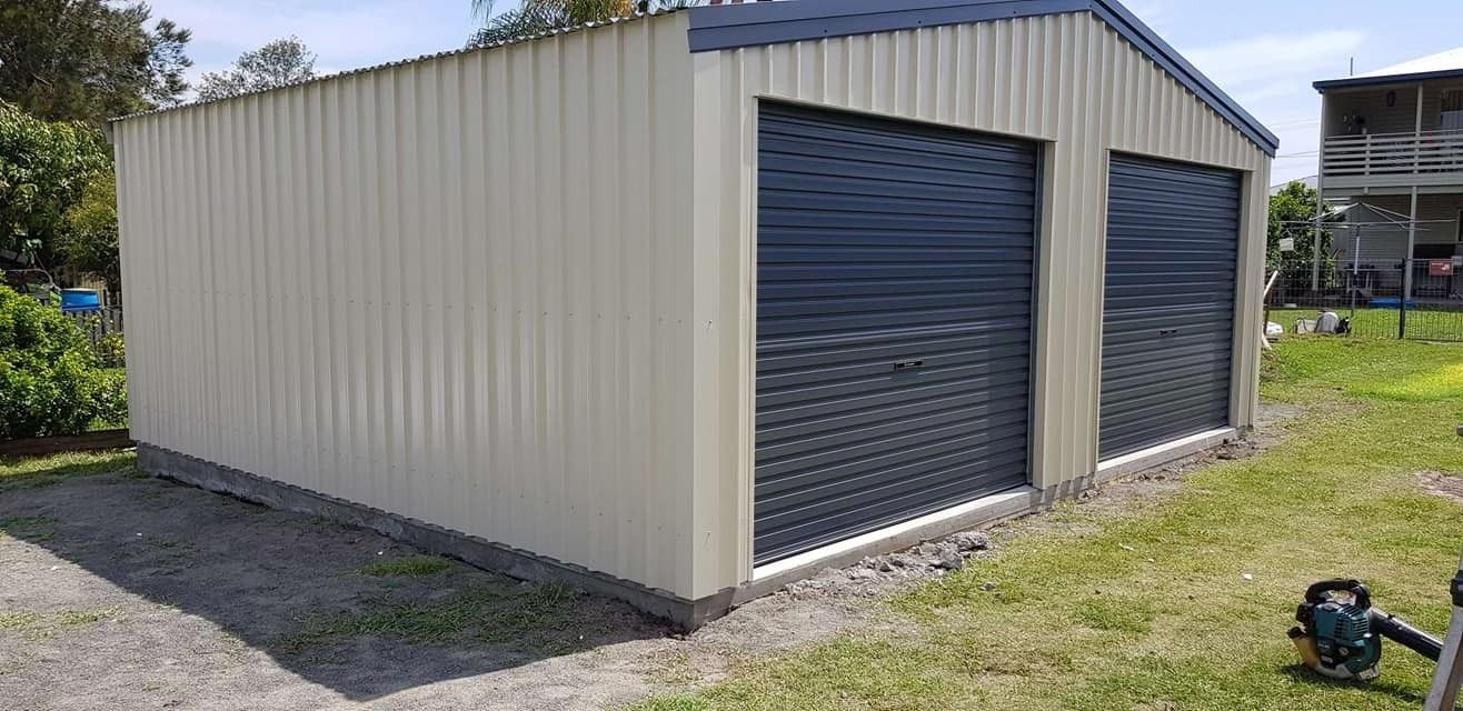A Two-bay Metal Shed With Dark Blue Roll-up Doors and a Light Beige Exterior Sits on a Concrete Foundation in a Grassy Yard — North Coast Sheds & Garages in Brunswick Heads, NSW