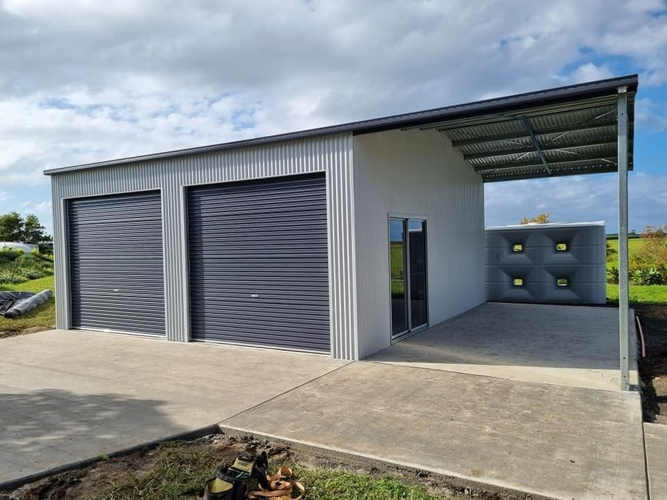 A Gray Metal Garage With Two Roll-up Doors, a Covered Side Area, and a Water Tank Sits on a Concrete Pad Under a Cloudy Sky — North Coast Sheds & Garages in Coraki, NSW