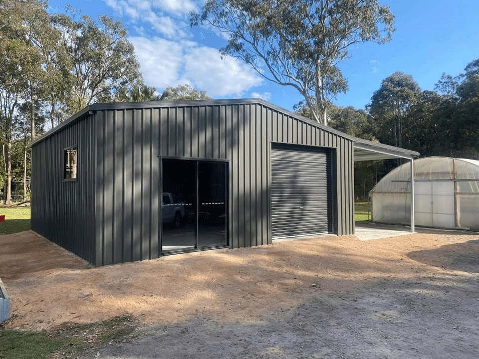 Dark Gray Metal Shed With Garage Door and Glass Doors, Situated on a Gravel Lot With Trees in the Background — North Coast Sheds & Garages in Coraki, NSW