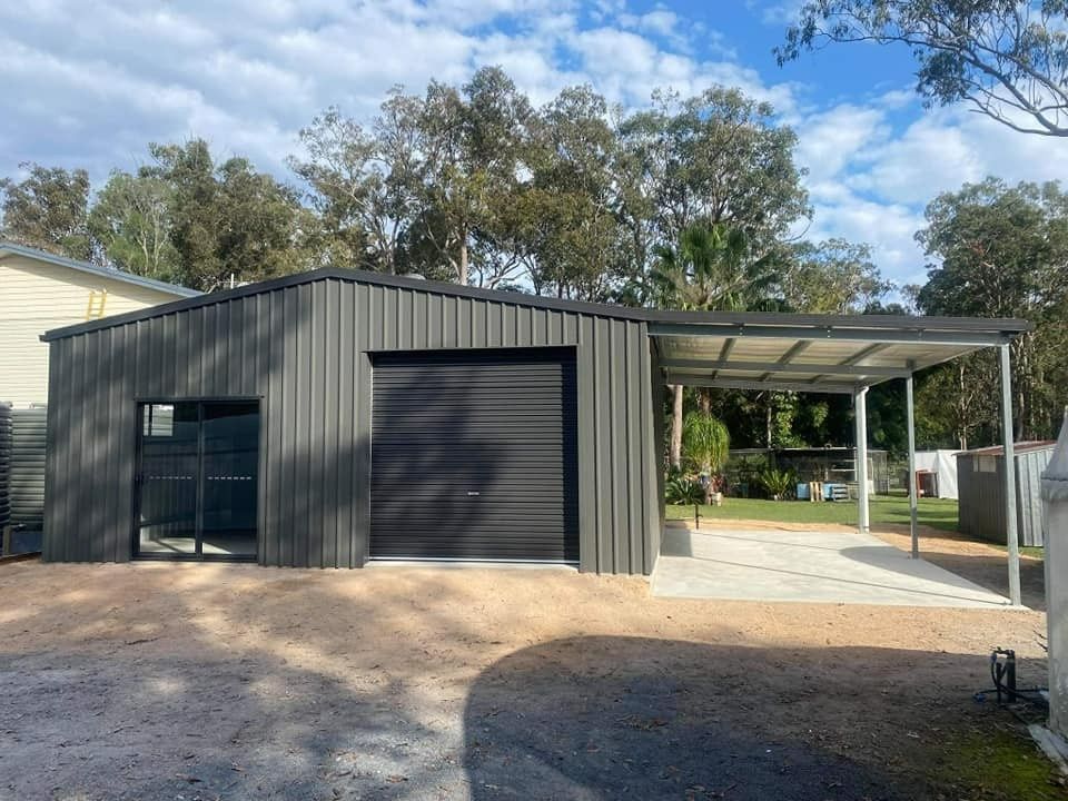 Dark Gray Metal Shed With a Roller Door, Adjacent Carport, and a Paved Area. Set in a Yard With Trees — North Coast Sheds & Garages in Coraki, NSW