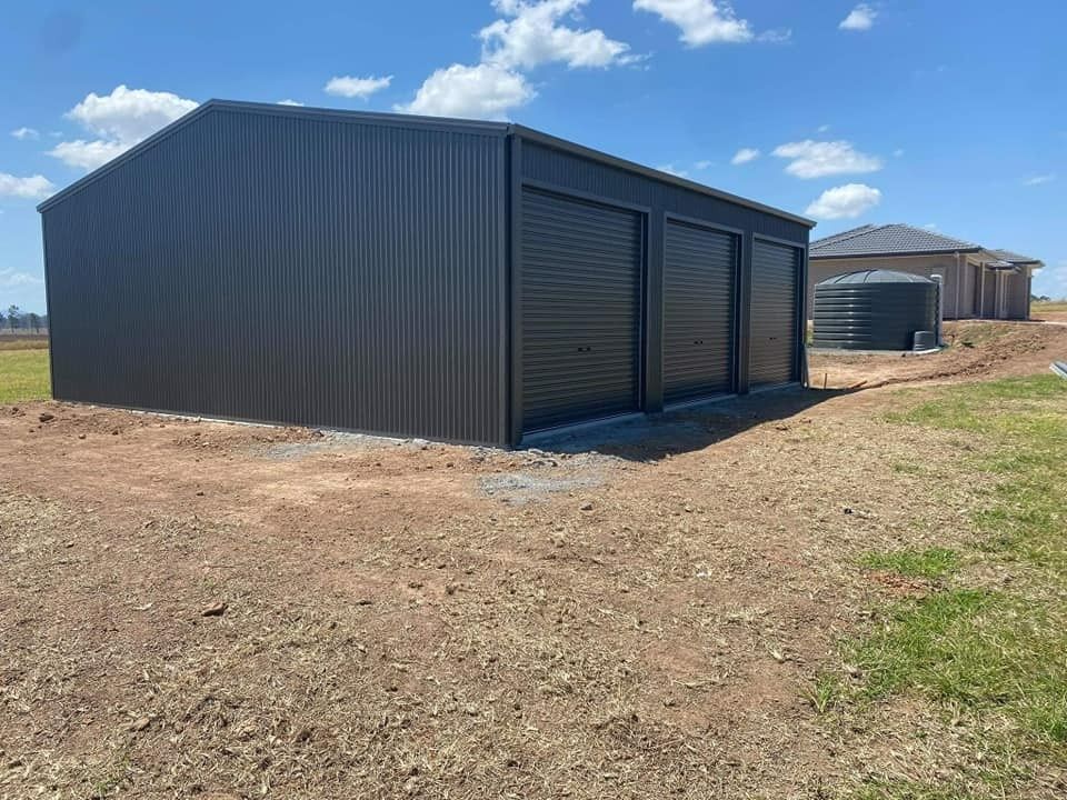 Dark Gray Metal Shed With Three Roller Doors on a Gravel Base, Situated on a Grassy Lot Under a Blue Sky — North Coast Sheds & Garages in Coraki, NSW