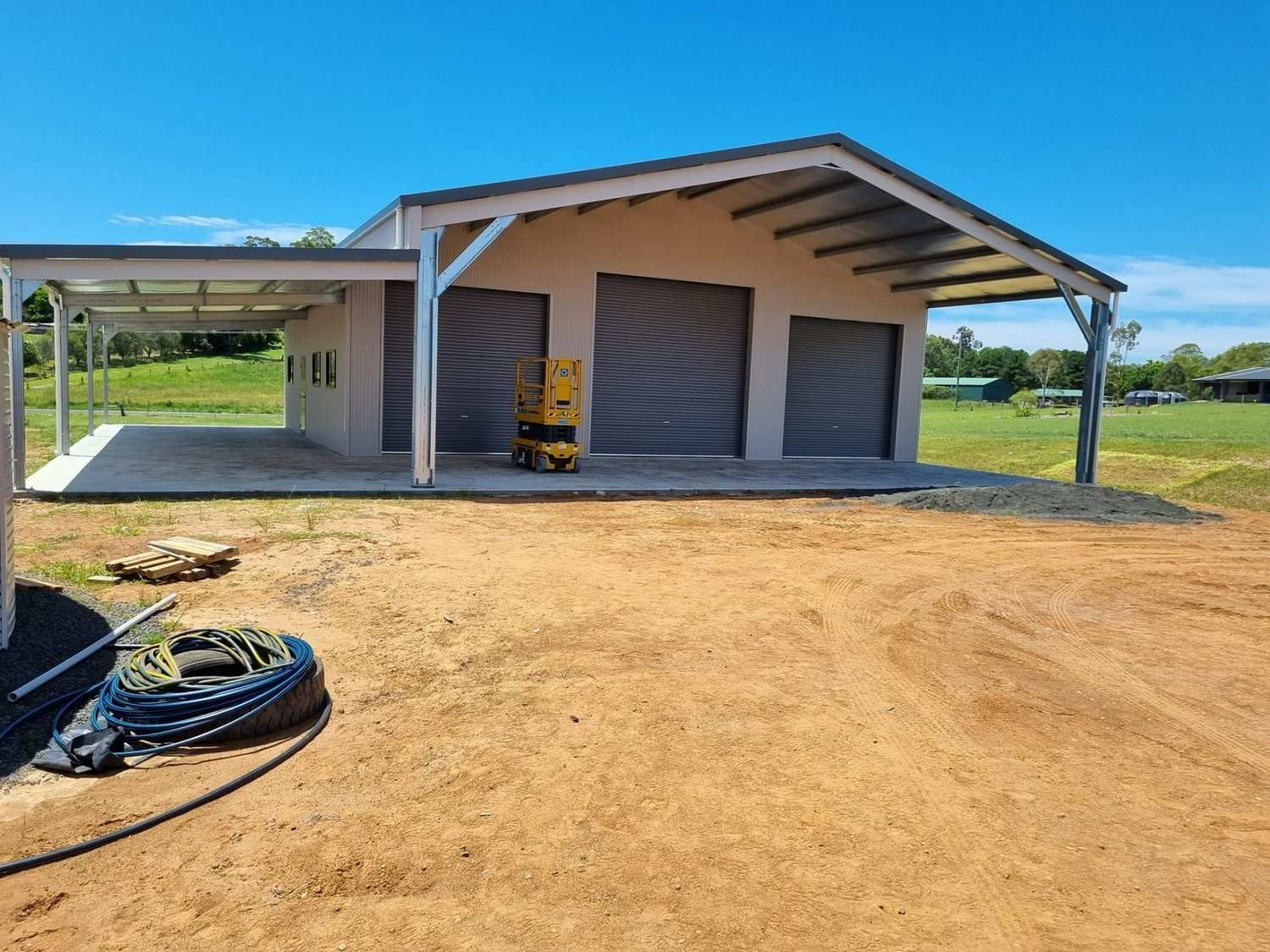 Tan Building With Three Garage Doors and a Carport, Set in a Dirt Lot With a Blue Sky in the Background — North Coast Sheds & Garages in Coraki, NSW