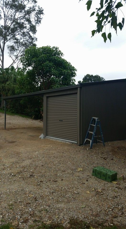 A Brown Shed With a Roll-up Door and Attached Carport on a Gravel Driveway. a Ladder and a Green Crate Sit Nearby — North Coast Sheds & Garages in Kyogle, NSW