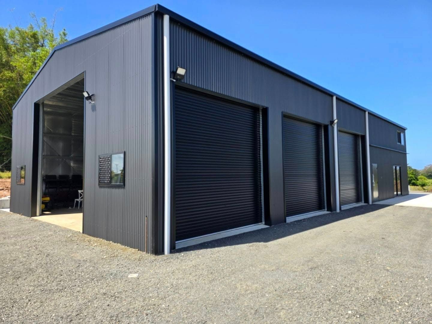 Dark Gray Metal Building With Multiple Roll-up Garage Doors on a Gravel Surface Under a Blue Sky — North Coast Sheds & Garages in Yamba, NSW