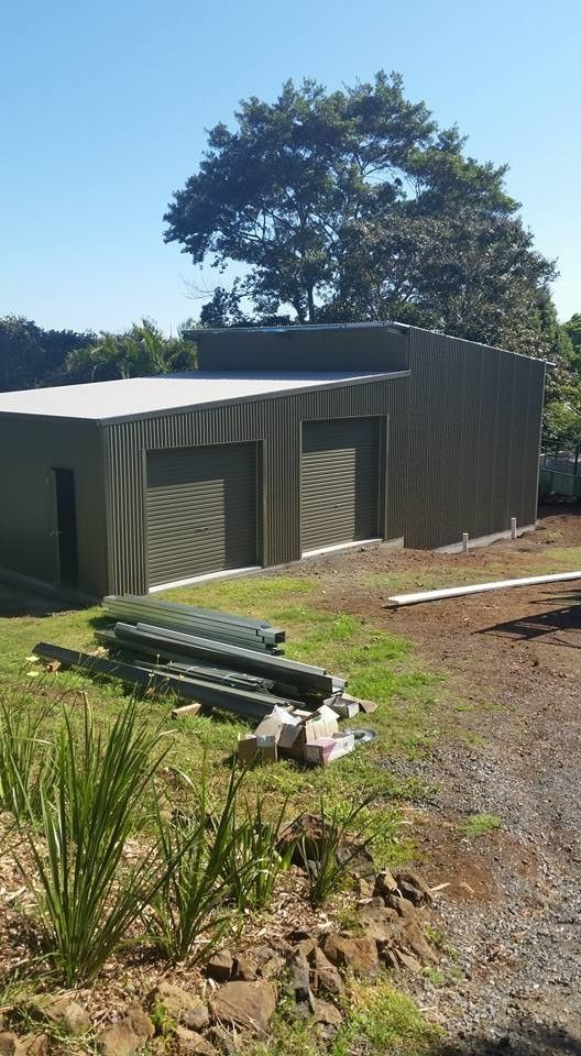 A Metal Garage With Two Doors and a Smaller Side Door Sits on a Grassy Area. Green Metal Beams Lay on the Ground — North Coast Sheds & Garages in Casino, NSW