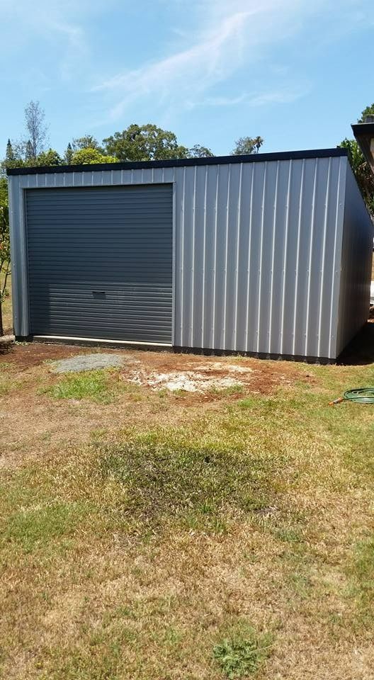 A Silver Metal Shed With a Gray Roller Door Stands on a Grassy Lawn Under a Bright Blue Sky — North Coast Sheds & Garages in Lismore, NSW