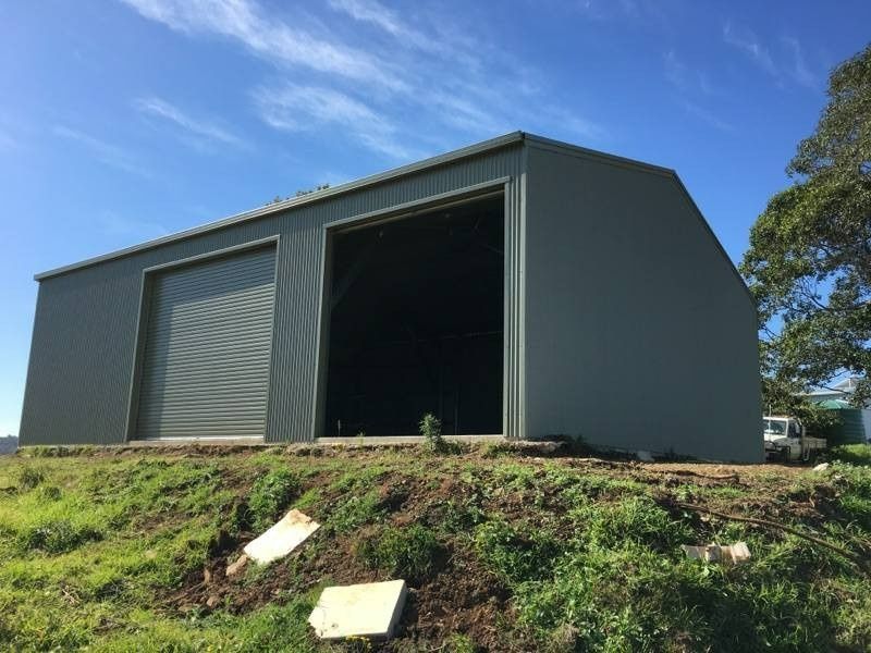 A Large, Green Metal Shed With Two Bays, One Open and One With a Closed Garage Door, Sits on a Grassy Hill Under a Blue Sky — North Coast Sheds & Garages in Bangalow, NSW