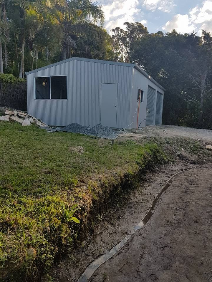 A Light Gray Metal Shed With a Window, Door, and Garage Bays Sits on a Grassy Hill in Front of Trees — North Coast Sheds & Garages in The Northern Rivers, NSW