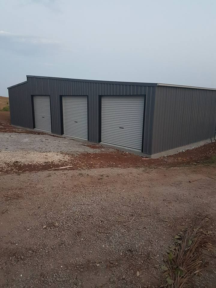 Gray Metal Building With Three Garage Doors in a Dirt Field Under a Cloudy Sky — North Coast Sheds & Garages in Byron Bay, NSW