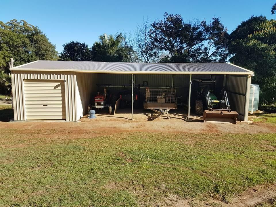 A Beige Metal Shed With a Closed Garage Door, Sheltering a Tractor, Trailer, and Other Farm Equipment. the Scene is Set in a Rural, Sunny Environment — North Coast Sheds & Garages in Bangalow, NSW