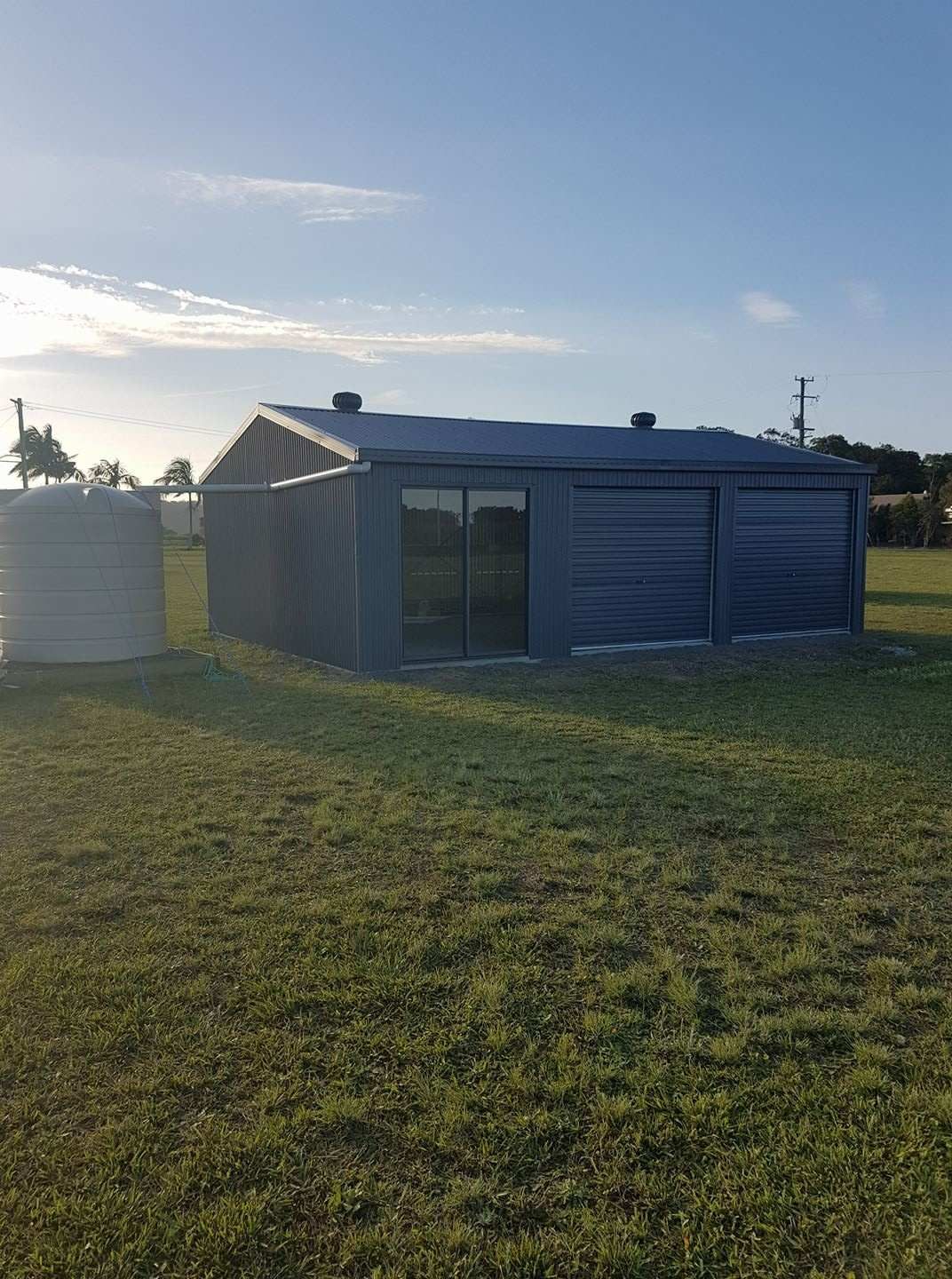 Blue Metal Shed With Large Glass Doors and Roller Doors in a Grassy Field; a Water Tank Stands to the Left — North Coast Sheds & Garages in Mullumbimby, NSW