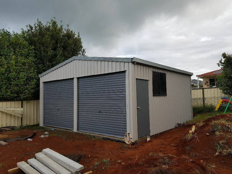A Two-car Garage With Gray Roll-up Doors and a Side Door and Window. the Structure is Metal and Sits on a Dirt Lot Under a Cloudy Sky — North Coast Sheds & Garages in Brunswick Heads, NSW