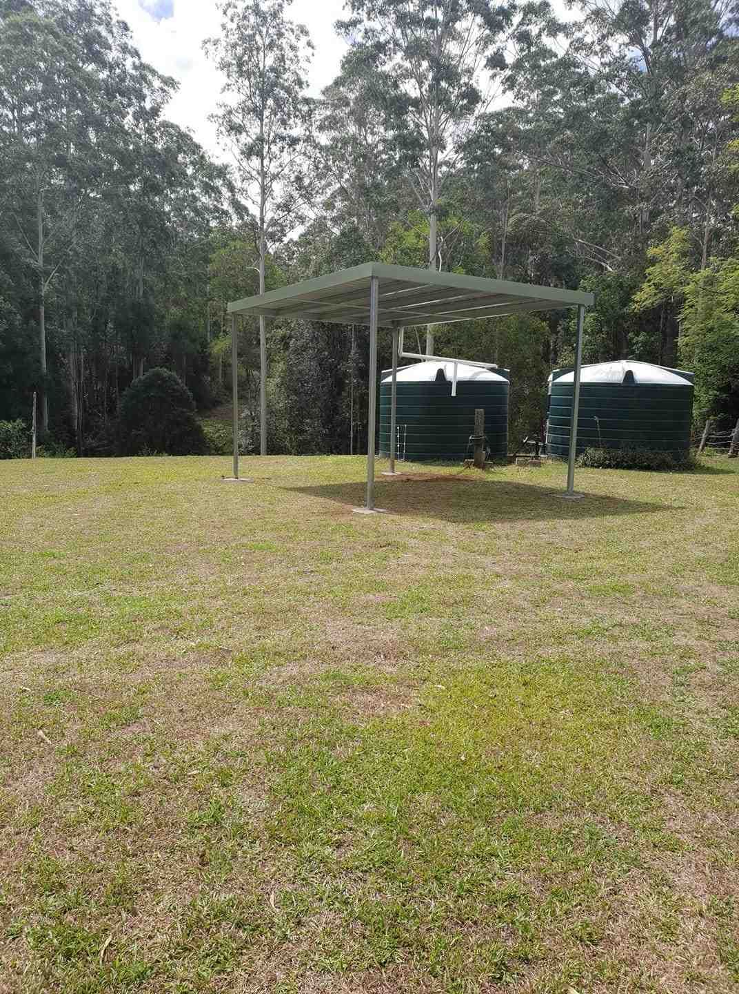 A Grassy Yard With a Metal Shelter Covering Two Green Water Tanks. Tall Trees Form a Backdrop — North Coast Sheds & Garages in Coraki, NSW