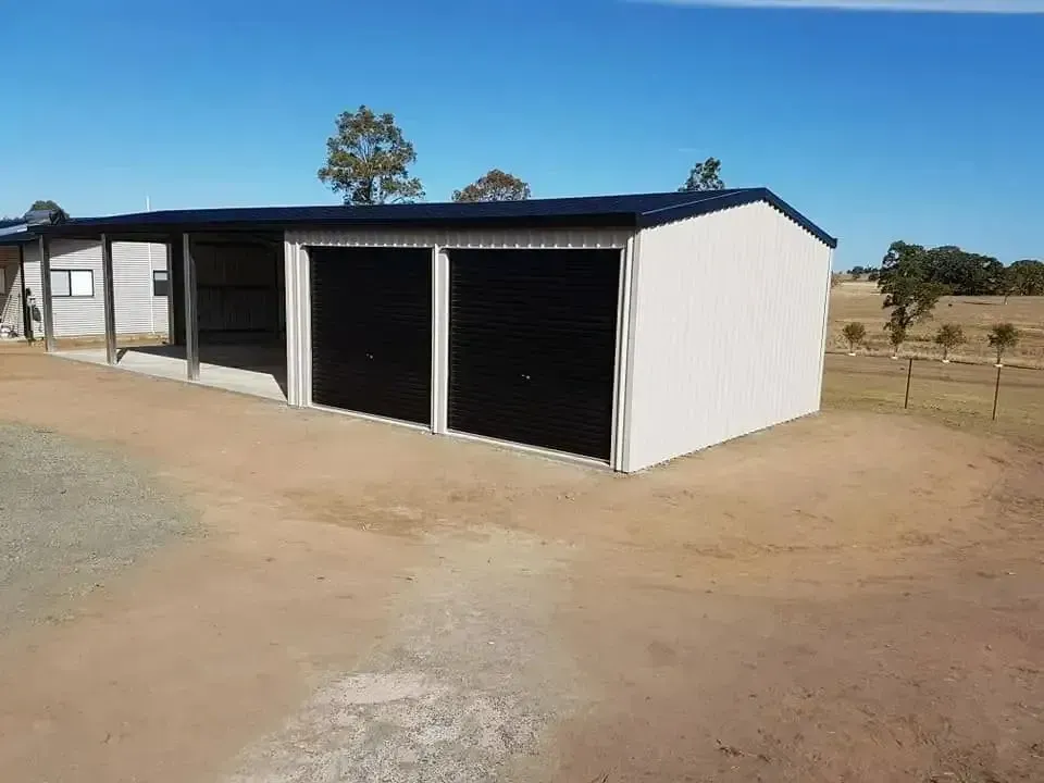 A Beige Metal Shed With Two Black Garage Doors and an Attached Carport on a Dirt Driveway Under a Blue Sky — North Coast Sheds & Garages in Grafton, NSW