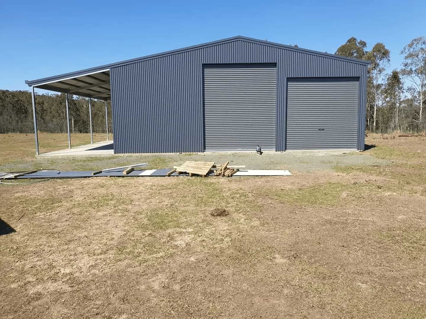 Blue Corrugated Metal Shed With Two Garage Doors and a Carport, Set in a Grassy Field — North Coast Sheds & Garages in Coraki, NSW